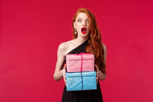 Celebration, holidays and women concept. Portrait of amazed and excited, ambushed young redhead female in black dress, drop jaw holding two cool gifts, stand red background.