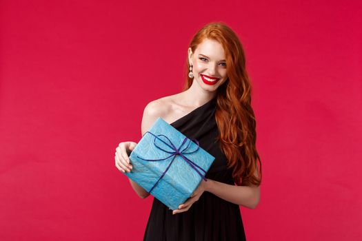 Celebration, holidays and women concept. Portrait of gorgeous young caucasian woman with ginger hair, wear red lipstick black evening dress, look camera happy smiling, holding birthday gift.