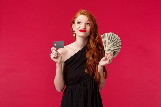 Luxury, beauty and money concept. Dreamy rich and lucky good-looking redhead woman thinking how waste her prize cash, smirk indecisive and looking up, put money on deposit credit card or shopping.