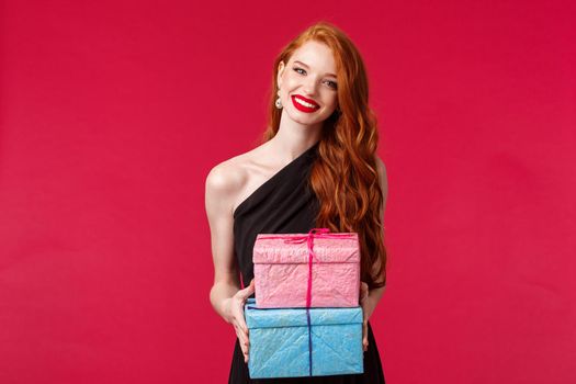 Celebration, holidays and women concept. Charming redhead female in black evening dress, red lipstick, give presents to birthday girl, party owner, holding two gifts and smiling cheerful.