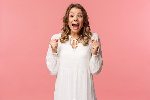 Portrait of surprised lucky hopeful, blond girl winning, hear great news and chanting, gasping open mouth smiling joyful, jumping from happiness and joy, standing pink background.