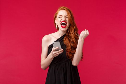 Portrait of successful and pleased, lucky redhead girl winning, receive great news via message, gift pump like champion, scream yes she did it, triumphing in black elegant dress, red background.