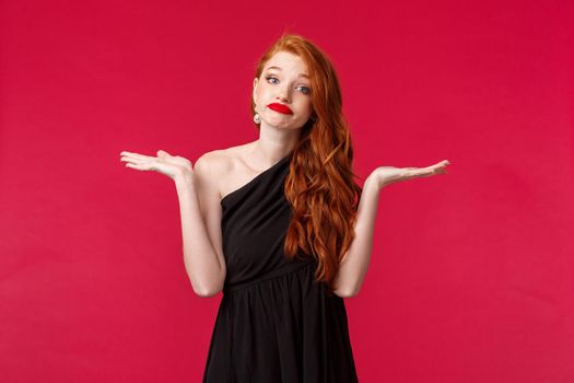 Portrait of careless indecisive young caucasian female redhead in elegant black dress, shrugging and smirk uncertain, have no idea, cant tell, dont know answer, standing black background.