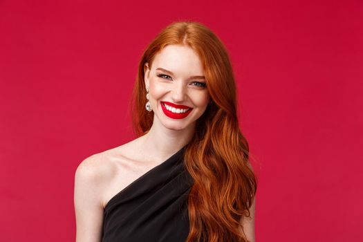 Romance, elegance, beauty and women concept. Elegant good-looking redhead woman in black stylish dress feeling hot and sassy, wear red lipstick, smiling confident, red background.