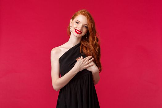 Fashion, luxury and beauty concept. Elegant good-looking redhead woman in black dress, with red lipstick and luxurious outfit, tilt head and smiling thankful, hold hand on heart touched with praises.