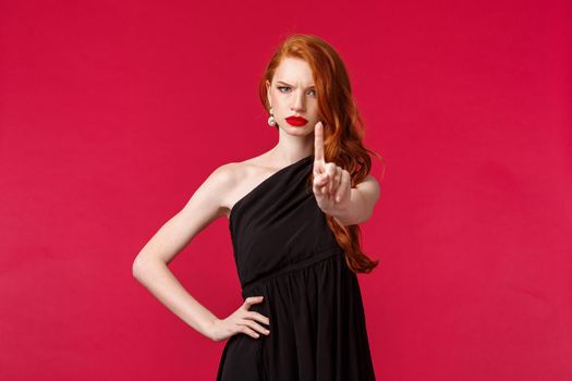 Fashion, luxury and beauty concept. Serious and displeased sassy redhead woman in luxurious stylish black dress, red lipstick, showing warning sign, shaking one finger scolding person bad decision.