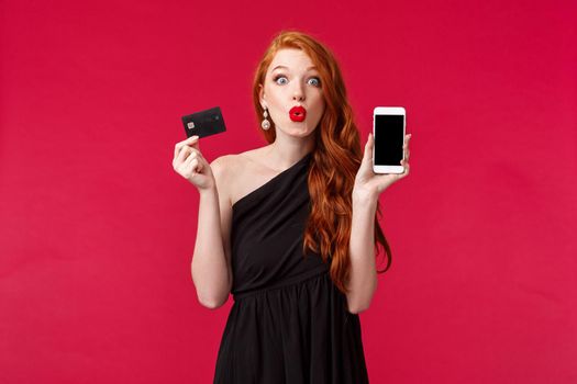 Fascinated and excited good-looking european woman with long curly ginger hair, wear black dress, holding mobile phone, showing smartphone display and credit card, shopping and internet concept.