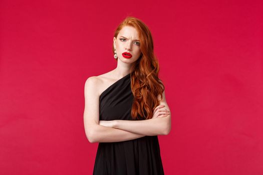 Portrait of serious and concerned, displeased young redhead woman in black dress, cross hands chest, frowning at camera wtih accusation and judgement, listening to something frustrating.