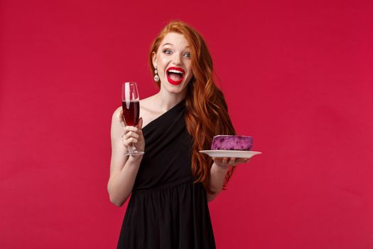 Portrait of gorgeous festive and excited woman celebrating birthday, feel amused and happy, holding glass champagne and b-day cake, having fun, wear black elegant dress over red background.