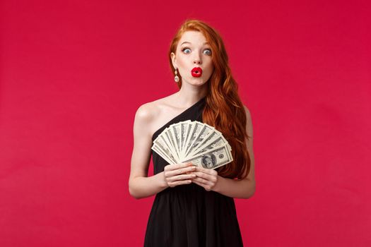 Luxury, beauty and money concept. Amazed silly good-looking young redhead woman in black stylish dress folding lips and looking surprised as winning cash, receive prize, stand red background.