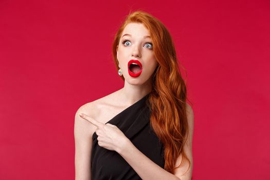 Close-up portrait of speechless impressed redhead woman drop jaw in amazement, seeing something stunning and cool, wear black dress, red lipstick, stare and pointing finger left, red background.