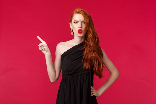 Beauty, fashion and women concept. Disappointed and dubious young redhead woman pointing finger left and frowning at camera upset, complain or being frustrated, wear black evening dress.