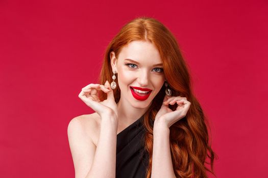 Elegance, beauty and fashion concept. Close-up portrait of sassy good-looking seductive young redhead woman touching her earrings and looking camera self-assured, flirting over red background.