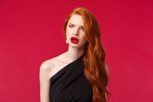 Close-up portrait of perplexed and confused young redhead woman seeing something strange, squinting and frowning as looking left troubled identify what is this, stand red background.