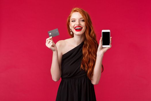 Portrait of gorgeous charismatic redhead woman in black dress, showing mobile phone display and credit card, paying for stunning black dress online in internet store, look satisfied smiling.