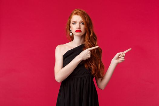 Celebration, events, fashion concept. Portrait of uneasy and sad pretty redhead woman in black dress, grimacing and frowning distressed, feel grief or regret, pointing fingers right, red background.