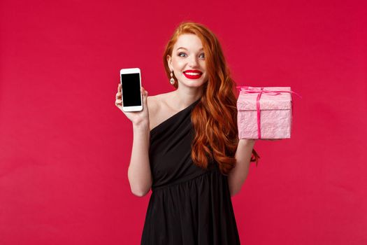 Portrait of excited and amused smiling, wondered redhead woman didnt expact receive gift so fast, showing mobile phone application, online delivery app, hold pink present over red background.