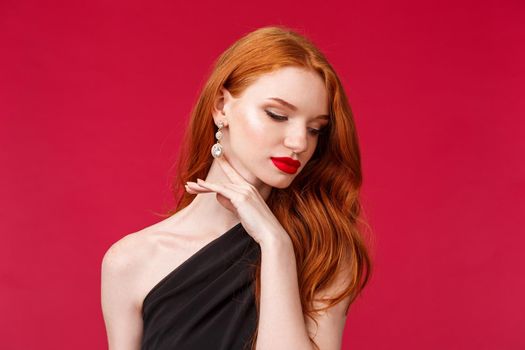 Romance, elegance, beauty and women concept. Close-up portrait of seductive, sensual and feminine redhead adult woman in black stylish dress, earrings and makeup, look away touching gently jawline.