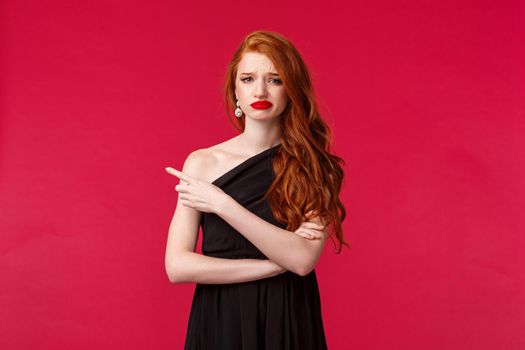 Portrait of skeptical, picky elegant redhead girlfriend in stylish black dress, grimacing disappointed, pointing finger left at something bad, dislike and disagree, stand red background.
