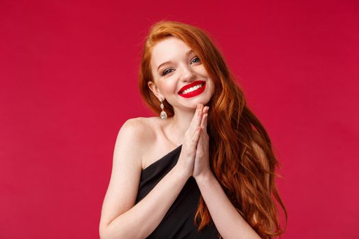 Close-up portrait of tender and feminine young pretty woman with ginger hair, black evening dress, clap hands look hopeful, begging for favour with bright beaming smile, red background.