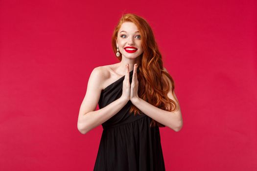 Portrait of hopeful and optimistic, excited redhead female in black dress, clap hands, applause thrilled watching stunning performance with amused smile, standing red background. Copy space