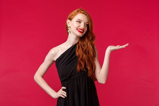 Portrait of luxurious good-looking redhead woman in black evening dress, attend event, look at blank space smiling, holding product on arm, promotion of company, red background.