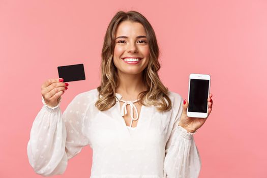 Finance, shopping and technology concept. Close-up portrait of cheerful, romantic blond cute girl in white dress, holding credit card and mobile phone, showing smartphone display, application promo.