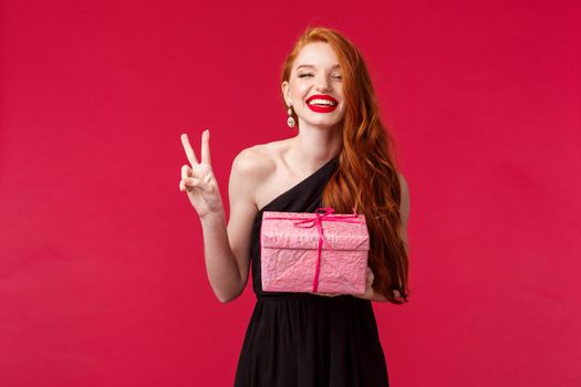 Celebration, holidays and women concept. Happy gorgeous young redhead female with long red hair, lipstick, wear black evening dress, have birthday party, laughing and smiling, hold gift.