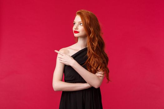 Celebration, events, fashion concept. Portrait of elegant young redhead slim woman in black stylish dress, wear makeup red lipstick, look and pointing left with curious interested face.