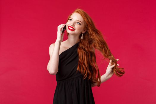 Portrait of gorgeous, elegant young redhead girl with long curly ginger hair, touching strand carefree talking on phone, receive praises during conversation, smiling look dreamy in black dress.