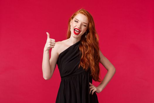 Fashion, luxury and beauty concept. Portrait of sassy good-looking elegant woman with red hair, black dress, wink at camera seductive, show thumb-up in like, approval gesture, red background.