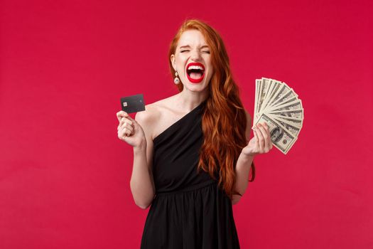 Luxury, beauty and money concept. Portrait of triumphing lucky attractive young redhead woman in black stylish dress screaming from delight and joy, winning prize cash, hold credit card and dollars.