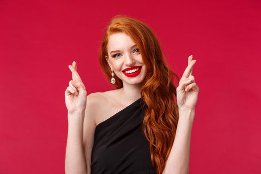 Close-up portrait of optimistic gorgeous redhead woman, praying for dream come true, cross fingers and smiling camera, assure she will win, determined receive prize, stand red background.