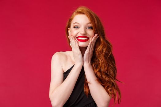 Close-up portrait of excited happy and surpirsed, cheerful gorgeous redhead woman with red lipstick, black dress, blushing happily touch cheeks and show beaming smile, red background.