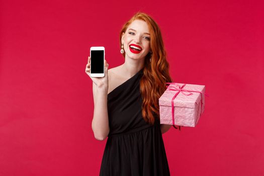 Portrait of gorgeous redhead woman in stylish black dress, laughing and smiling, red lipstick, showing mobile phone application, online show in smartphone display, hold gift box, red background.