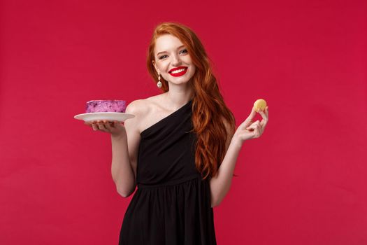Elegant young woman in stylish black dress, red lipstick wearing black trendy dress over red background, holding biscuit and cake, smiling camera, cant resist temptation of desserts.