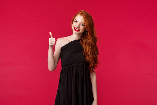 Elegance, fashion and woman concept. Portrait of satisfied charming young elegant redhead woman in black stylish dress over red background, show thumbs-up in approval or like sign.