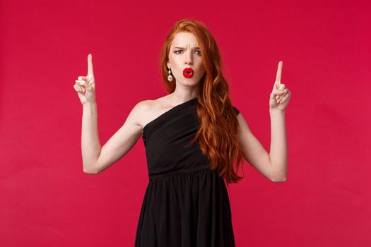 Celebration, events, fashion concept. Portrait of frustrated and puzzled redhead woman in red lipstick, black elegant dress, frowning disappointed, pointing up at something bad.