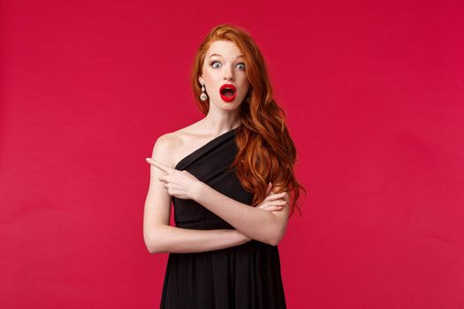 Elegance, fashion and woman concept. Portrait of ambushed and surprised young redhead woman, girl in black dress looking shocked, gossiping pointing finger left, red background.