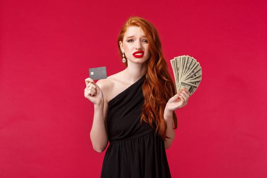 Luxury, beauty and money concept. Indecisive and unsure good-looking young redhead girlfriend holding lots of cash dollars and credit card, grimacing uncertain have doubts how waste it.