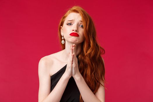 Pretty please. Hopeful cute silly redhead woman with red lipstick, black evening dress, asking eager to go party with girlfriends, hold hands in pray begging for something badly, red background.