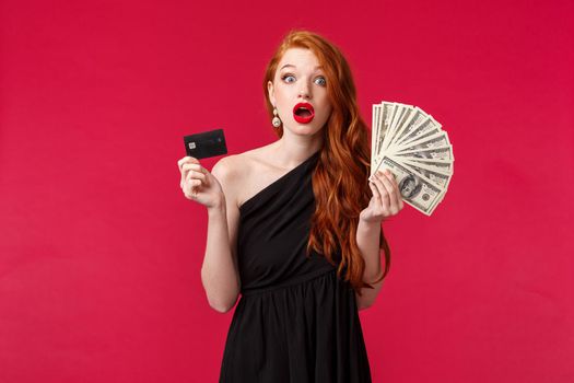 Luxury, beauty and money concept. Amazed silly young attractive woman suddenly got rich, winning prize cash, hold dollars and credit card, gasping and staring camera disbelief, red background.