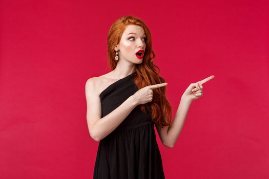Celebration, events, fashion concept. Surprised elegant and pretty redhead woman, ginger hair, wear beautiful black evening dress, looking and pointing right, turn attention at interesting promo.