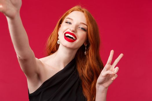 Close-up portrait of happy charming redhead woman enjoying party, taking selfie with peace sign and beaming smile, wear black evening dress and red lipstick, stand red background.