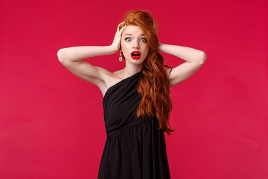 Portrait of shocked and ambushed young redhead female in elegant black dress, gasping nervously, look alarmed camera hold hands on head, standing startled over red background.