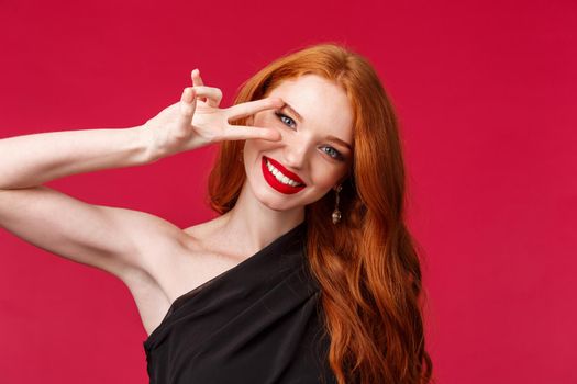 Elegance, beauty and fashion concept. Close-up portrait of gorgeous seductive redhead woman, caucasian female model in black dress, makeup and red lipstick, show peace sign near eye, smiling.