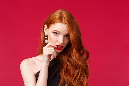 Close-up portrait of silly pretty feminine redhead woman with earrings, red lipstick and black dress, look from under forehead flirty, seducing someone acting clueless and dumb, red background.