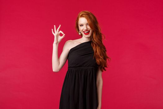 Elegance, fashion and woman concept. Portrait of sexy redhead assertive young woman in stylish elegant black dress, slim female model wearing prom outfit, show okay, excellent gesture smile pleased.