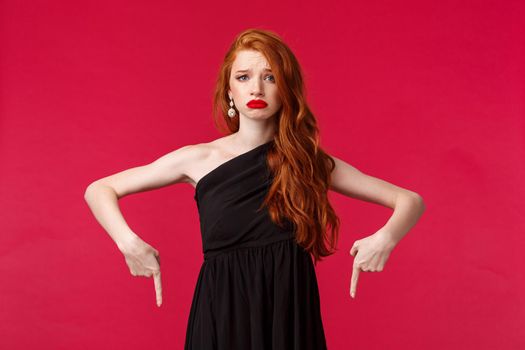 Portrait of gloomy and upset, grieving redhead woman in black dress, frowning unhappy looking camera with regret or uneasy emotions, pointing fingers down, stand red background.