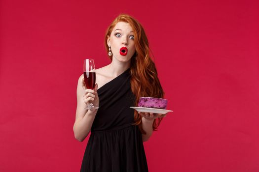 Portrait of amused and wondered, speechless cute redhead woman in stylish black dress, holding glass champagne and cake, folding lips surprised look camera, stand red background.